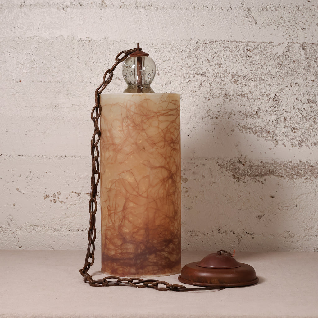 A vintage-style table lamp with a cylindrical amber-brown marbled stone or resin column, a dark metal chain, a clear glass globe shade, and a matching round base, set against a distressed off-white wall.