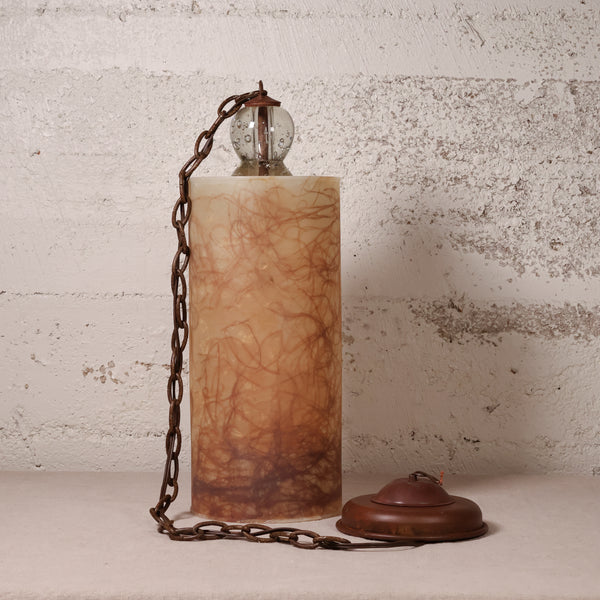 A vintage-style table lamp with a cylindrical amber-brown marbled stone or resin column, a dark metal chain, a clear glass globe shade, and a matching round base, set against a distressed off-white wall.