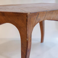 A close-up shot of a rustic wooden table showing its detailed grain, joint construction, and curved legs against a plain light background.
