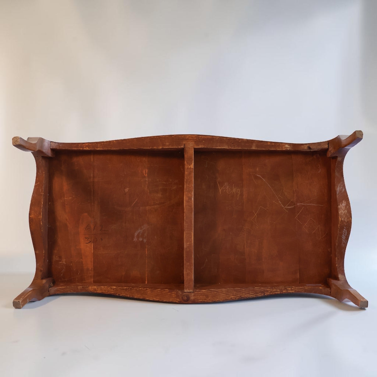 The underside of a rustic wooden table or bench, showing a warm brown wood grain, curved apron, and four short feet, photographed against a neutral light gray background.