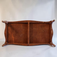 The underside of a rustic wooden table or bench, showing a warm brown wood grain, curved apron, and four short feet, photographed against a neutral light gray background.