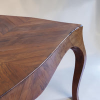 Close-up of a wooden table corner showing a curved cabriole-style leg and a herringbone wood top in warm medium brown tones.