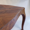 Close-up of a wooden table corner showing a curved cabriole-style leg and a herringbone wood top in warm medium brown tones.