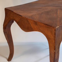 A close-up of a wooden table leg and corner, showing a warm medium-brown wood grain finish and a curved cabriole-style leg against a plain light background.
