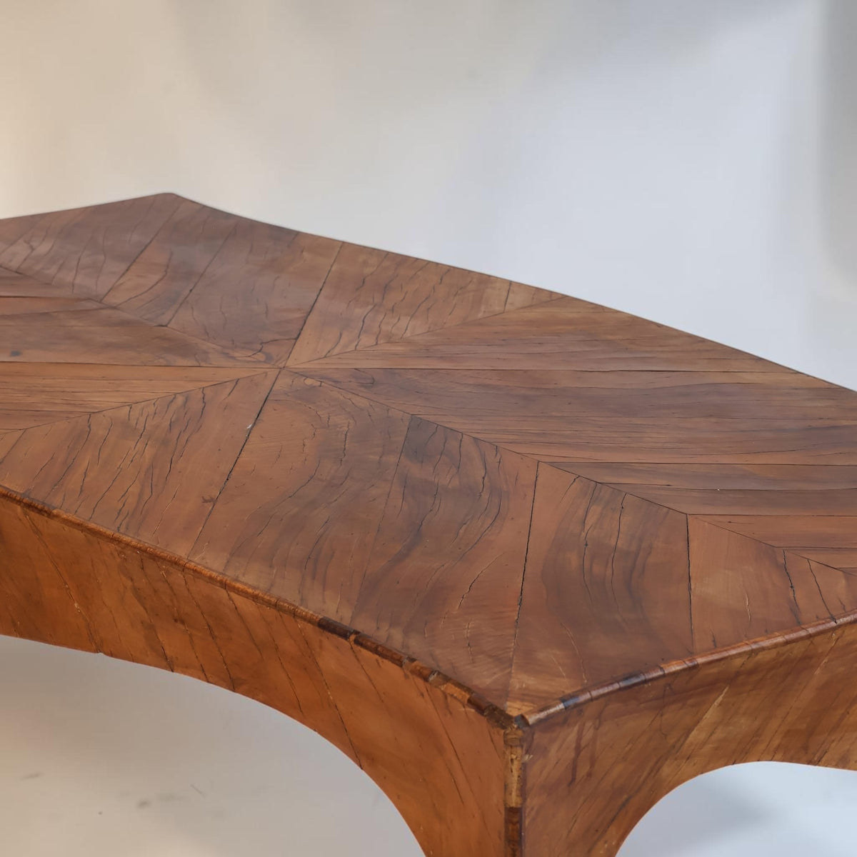 A close-up of a wooden table top with a curved apron and a geometric inlay pattern, showing rich warm brown wood grain against a plain light background.