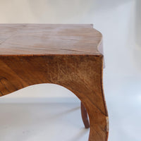 A close-up of a wooden table corner showing the curved apron, grain pattern, and tabletop surface against a plain light background.