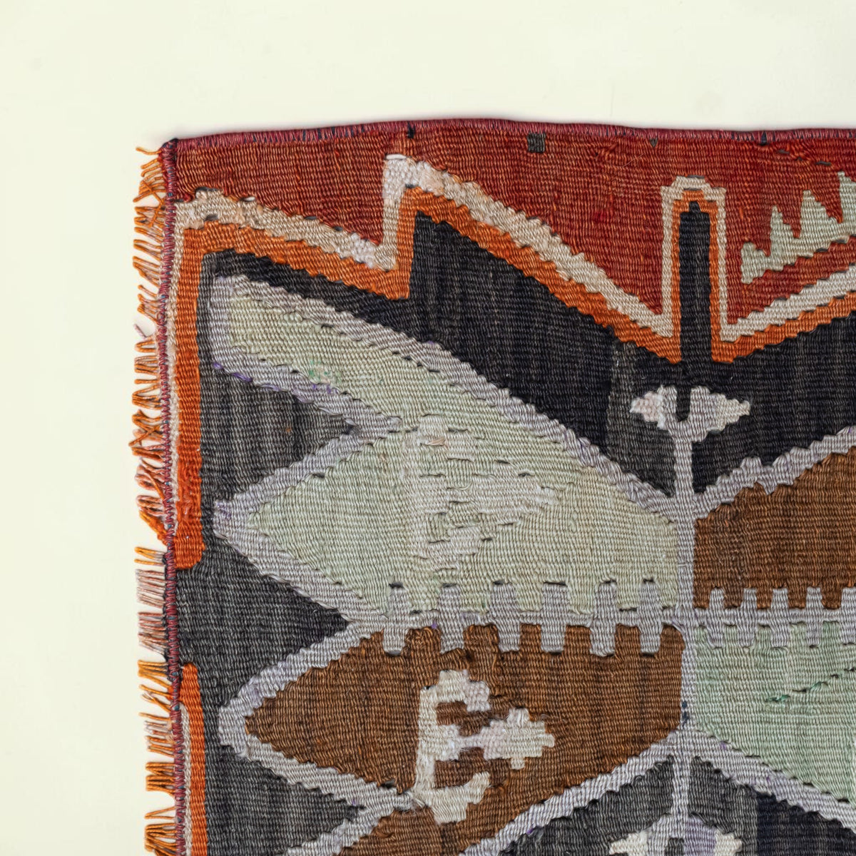 Close-up of a woven rug corner showing a geometric pattern in rust red, orange, charcoal gray, brown, pale green, and off-white, with fringe along the left edge on a light cream background.
