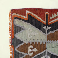 Close-up of a woven rug corner showing a geometric pattern in rust red, orange, charcoal gray, brown, pale green, and off-white, with fringe along the left edge on a light cream background.