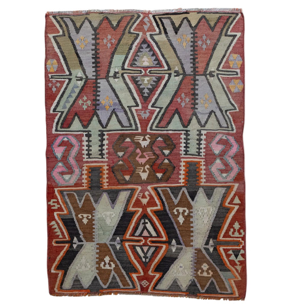 A rectangular kilim rug with a geometric tribal pattern in muted red, brown, gray, black, and pink, laid flat against a light beige background.