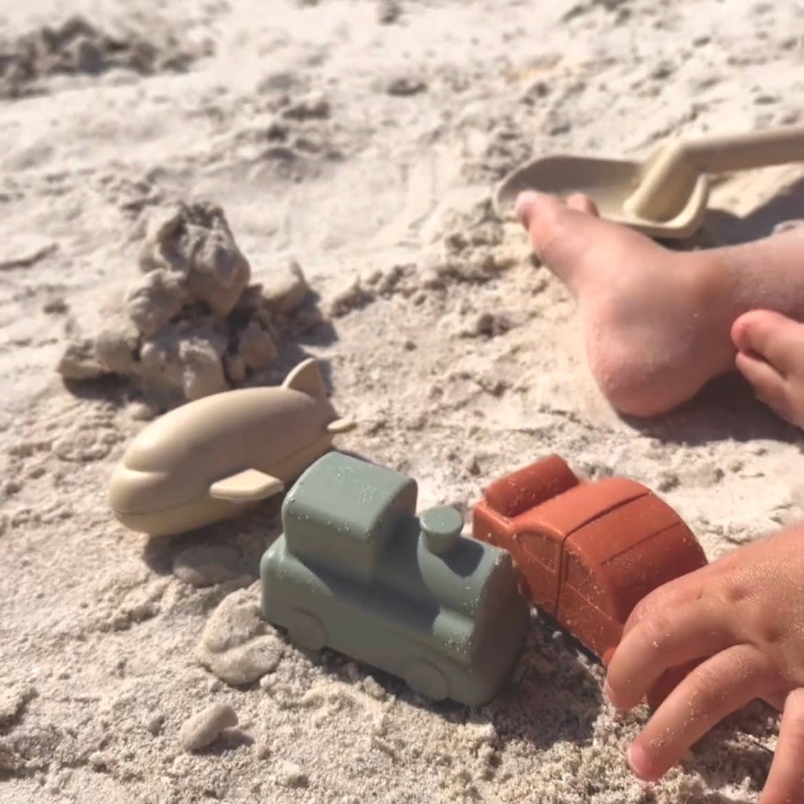 Children's toy cars on a sandy beach with a child's foot nearby.