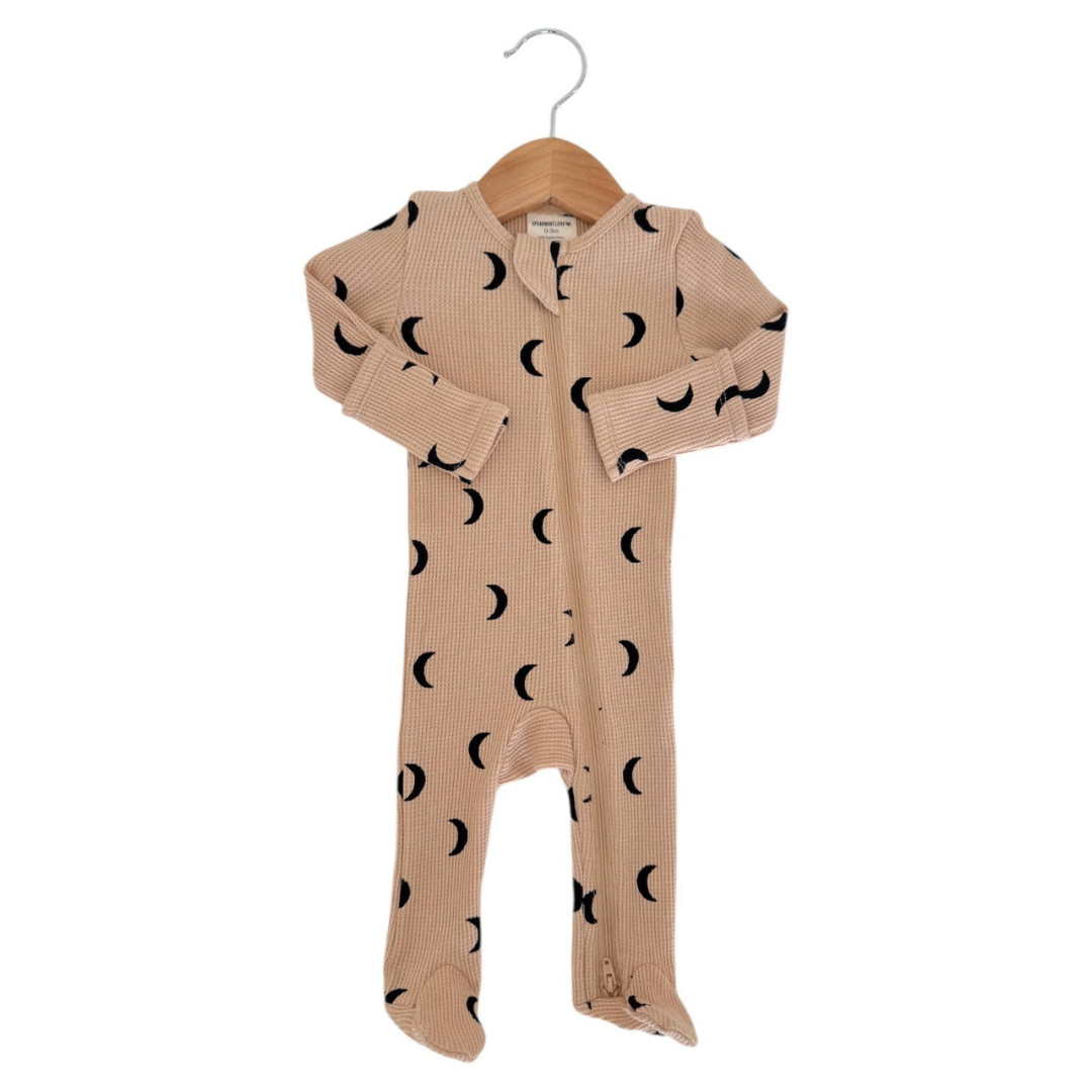 A baby footed one-piece sleeper in beige with a black crescent moon pattern, hanging on a wooden hanger against a white background.