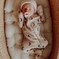Newborn baby in a beige outfit with black patterns, wearing a matching hat and holding a pacifier, lying in a woven basket.