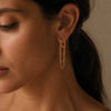 A close-up of a woman's ear wearing a delicate gold dangle earring with three layered chain loops, set against a warm neutral background.
