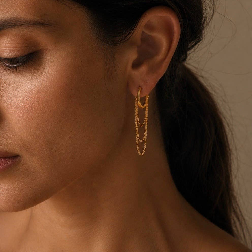 A close-up of a woman's ear wearing a delicate gold dangle earring with three layered chain loops, set against a warm neutral background.