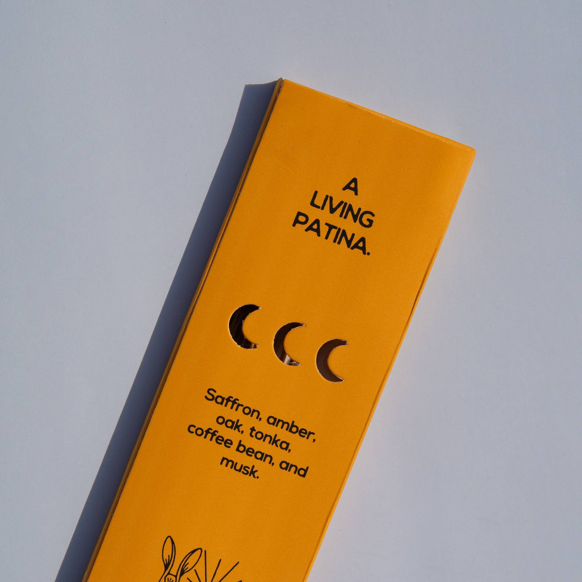 A tall, narrow yellow-orange package or box photographed on a light gray background. The front has black text reading 'A LIVING PATINA.
