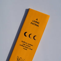 A tall, narrow yellow-orange package or box photographed on a light gray background. The front has black text reading 'A LIVING PATINA.
