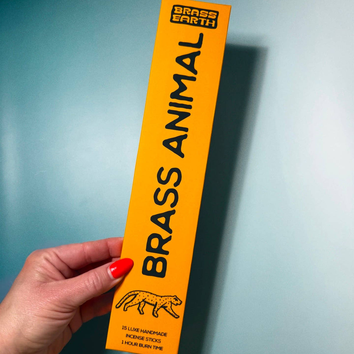 Hand holding a yellow 'Brass Animal' product packaging against a blue background
