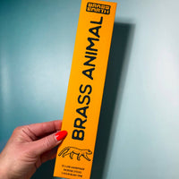 Hand holding a yellow 'Brass Animal' product packaging against a blue background