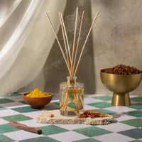 A reed diffuser with natural wood reeds in a clear glass bottle, styled on a green and white checkered tile surface with bowls of spices and incense sticks in a warm, rustic setting.