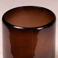 A close-up of a glossy amber brown glass container or vase against a light beige background.