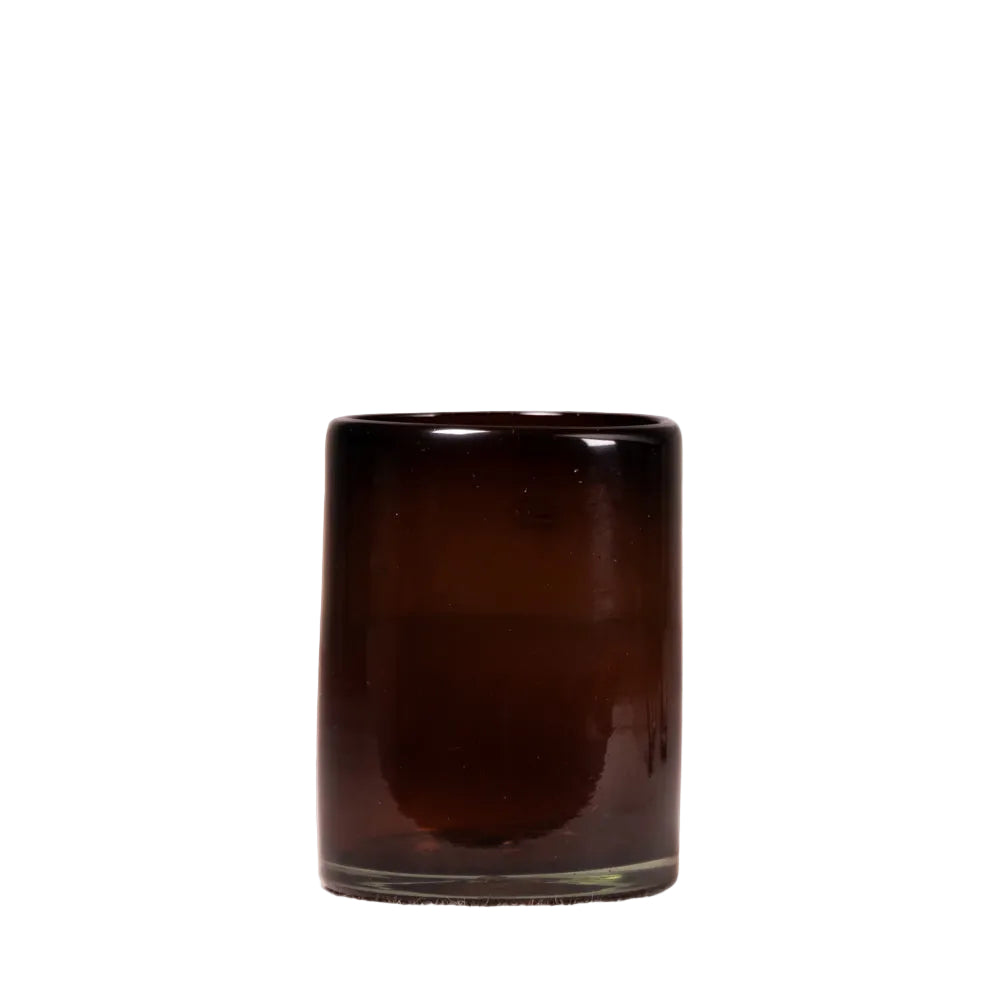 A small cylindrical dark amber glass candle holder or tumbler with a smooth, glossy finish, standing on a light beige surface against a textured, light off-white wall.