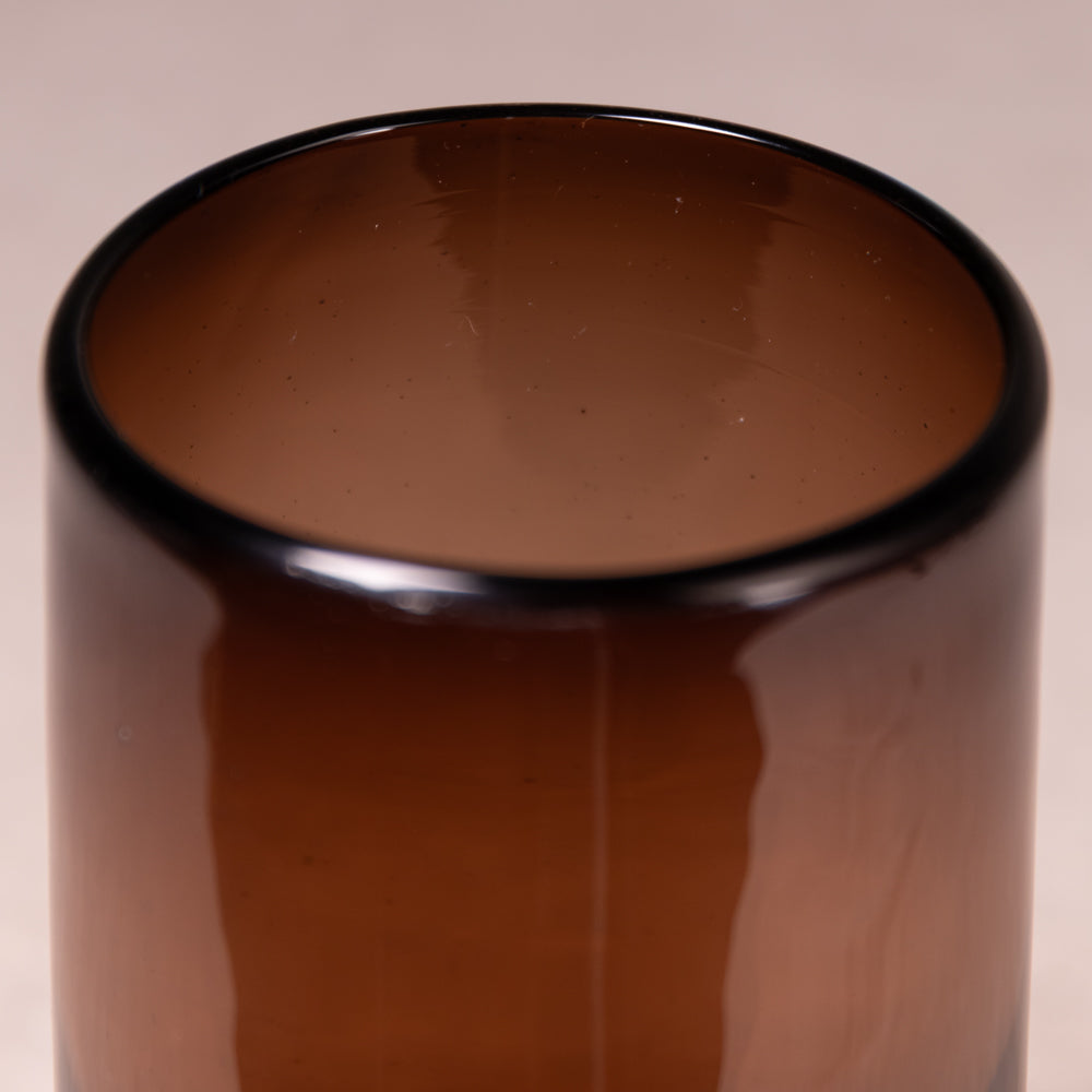 A close-up of a translucent amber brown glass tumbler or cup, showing its smooth glossy surface and dark rim against a neutral beige background.