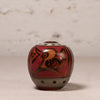A small hand-painted ceramic pot with a lid, featuring a red background, a brown bird design, and gray accents, set against a neutral textured backdrop.