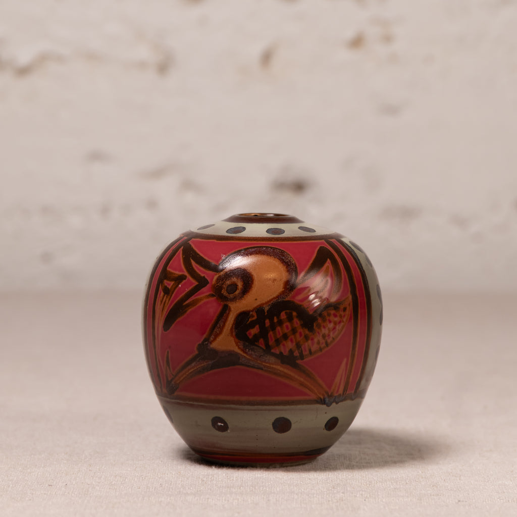 A small hand-painted ceramic pot with a lid, featuring a red background, a brown bird design, and gray accents, set against a neutral textured backdrop.