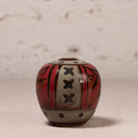A small hand-painted ceramic pot with a rounded body, featuring a red, black, and beige geometric pattern, set against a neutral textured background.