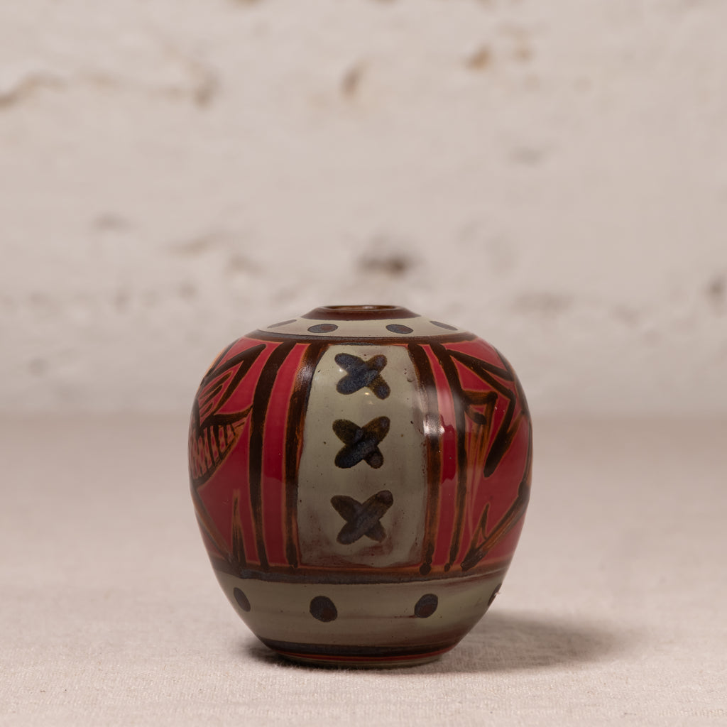 A small hand-painted ceramic pot with a rounded body, featuring a red, black, and beige geometric pattern, set against a neutral textured background.