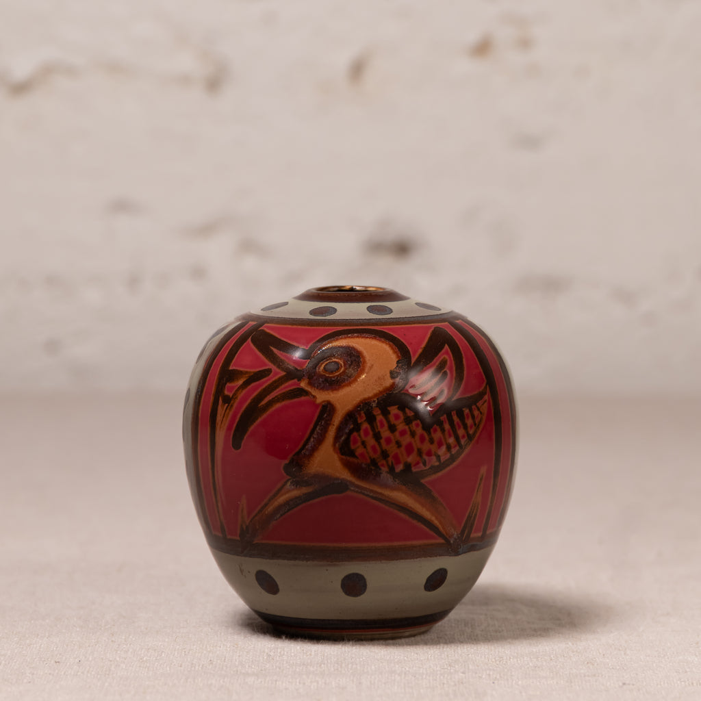 A small hand-painted ceramic pot with a lid, featuring a red and brown bird design with black accents, set against a neutral textured background.