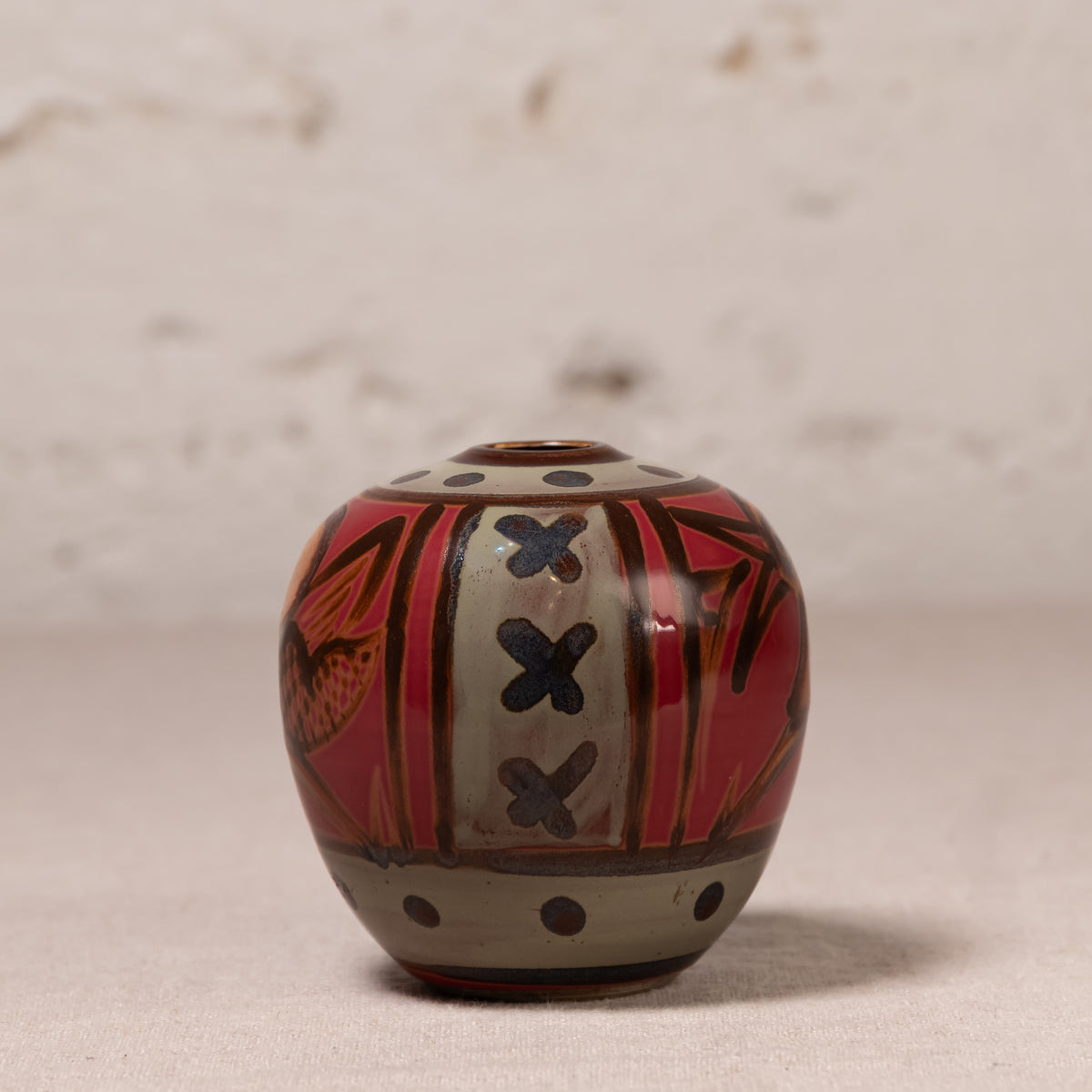 A small hand-painted ceramic pot with a rounded body, featuring a red and beige pattern with black accents, set against a neutral textured background.