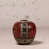 A small hand-painted ceramic pot with a rounded body, featuring a red and beige pattern with black accents, set against a neutral textured background.