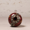 A decorative glass bead with a hand-painted pattern in red, orange, black, and beige, standing upright on a neutral surface against a textured light background.