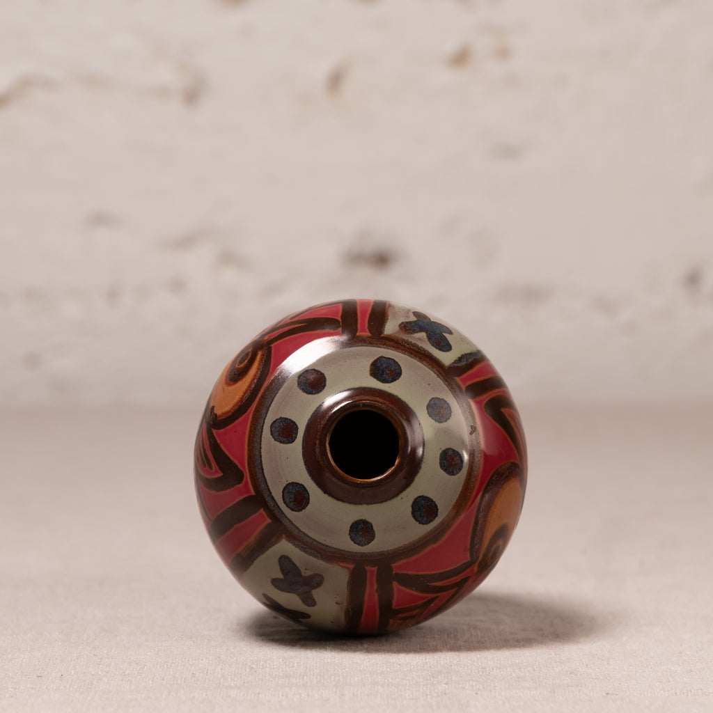 A decorative glass bead with a hand-painted pattern in red, orange, black, and beige, standing upright on a neutral surface against a textured light background.