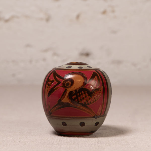 A small hand-painted ceramic pot with a lid, featuring a red background, a brown bird design, and gray accents, set against a neutral textured backdrop.