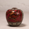 A small round ceramic vase with a lid, decorated with a stylized bird motif in brown and gold on a red background, with beige bands and dark polka dots.