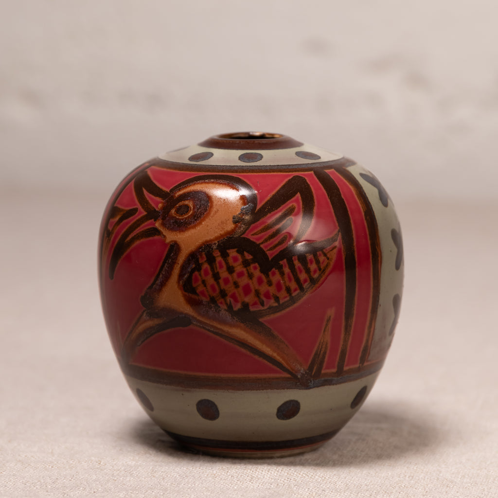A small round ceramic vase with a lid, decorated with a stylized bird motif in brown and gold on a red background, with beige bands and dark polka dots.