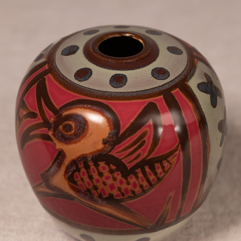 A round ceramic vase with a glossy finish, featuring a stylized bird design in brown and orange on a red background, with a cream-colored top band decorated with dark blue dots.
