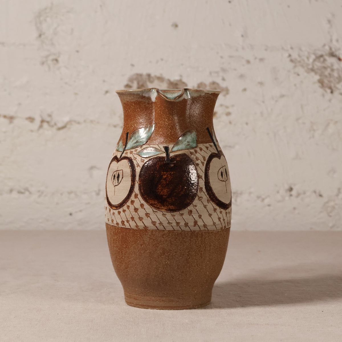 A handmade ceramic vase with a brown and beige speckled glaze, decorated with painted apple motifs and a patterned band, set against a textured off-white wall.