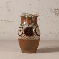 A handmade ceramic vase with a brown and beige speckled glaze, decorated with painted apple motifs and a patterned band, set against a textured off-white wall.