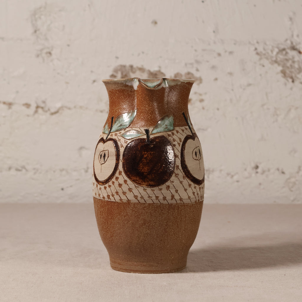 A handmade ceramic vase with a brown and beige speckled glaze, decorated with painted apple motifs and a patterned band, set against a textured off-white wall.