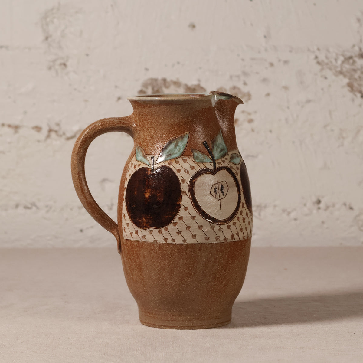 A handmade brown ceramic pitcher with a handle, featuring painted apple motifs and a textured cream band around the middle, set against a rustic white brick wall background.