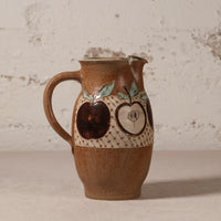 A handmade brown ceramic pitcher with a handle, featuring painted apple motifs and a textured cream band around the middle, set against a rustic white brick wall background.