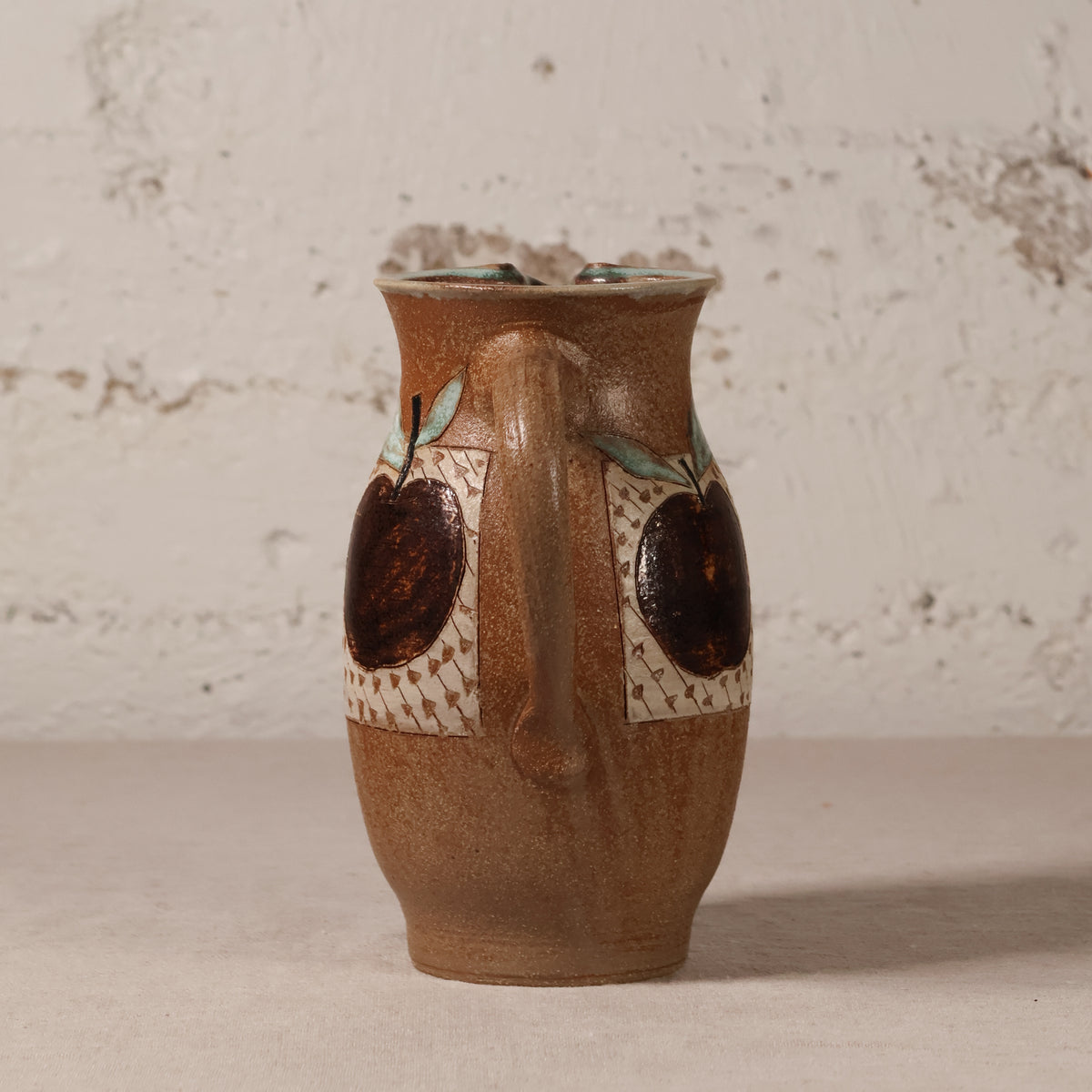 A handmade brown ceramic pitcher with a handle, decorated with two dark brown apple motifs and light green leaf accents on a textured beige patterned background, set against a rustic white plaster wall.
