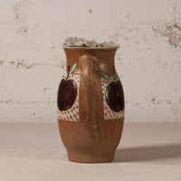 A handmade brown ceramic pitcher with a handle, decorated with two dark brown apple motifs and light green leaf accents on a textured beige patterned background, set against a rustic white plaster wall.
