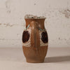 A handmade brown ceramic pitcher with a handle, decorated with two dark brown apple motifs and light green leaf accents on a textured beige patterned background, set against a rustic white plaster wall.