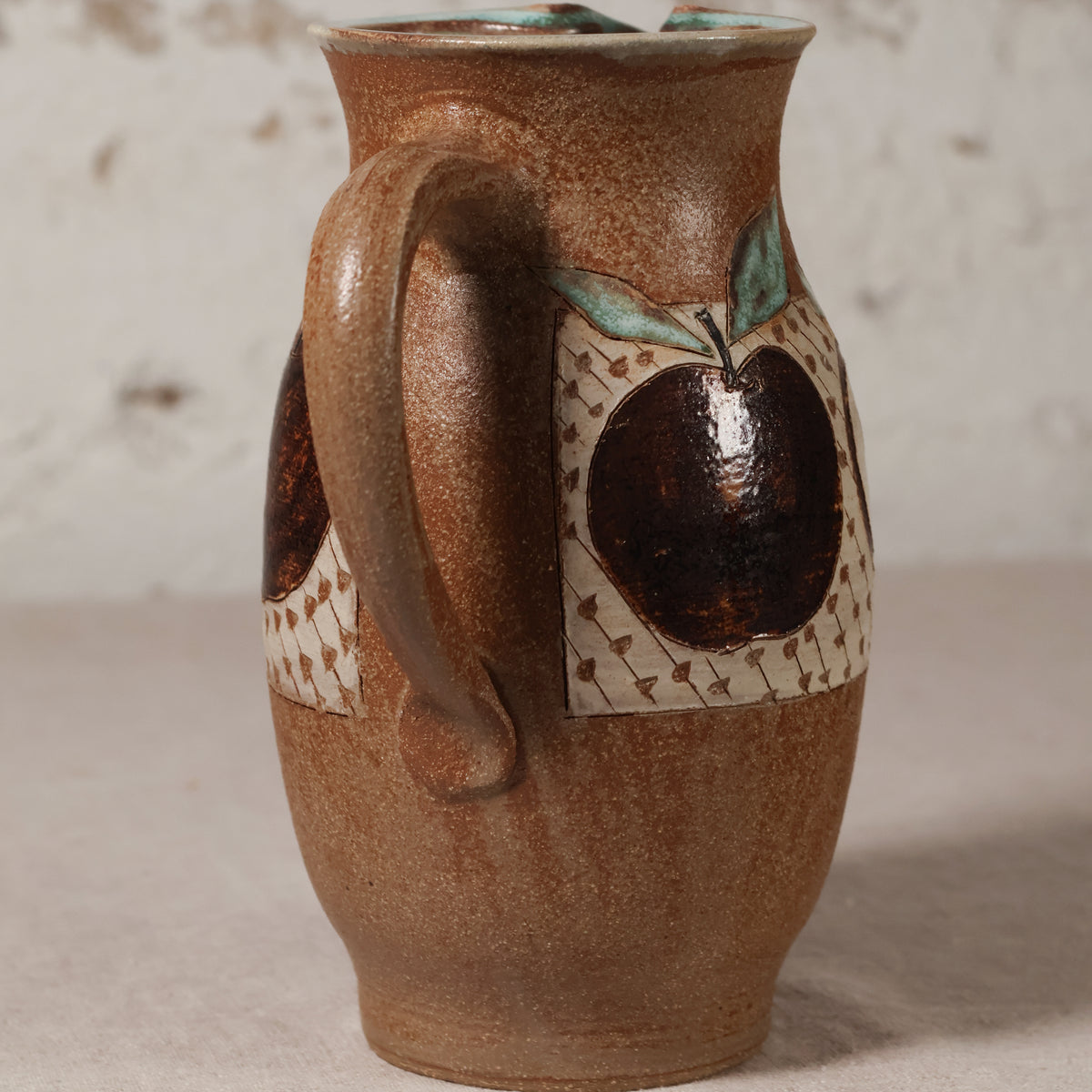 A handmade ceramic pitcher with a rounded brown body, a large handle, and a decorative pattern featuring dark brown apples with green leaves on a cream background.