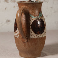 A handmade ceramic pitcher with a rounded brown body, a large handle, and a decorative pattern featuring dark brown apples with green leaves on a cream background.