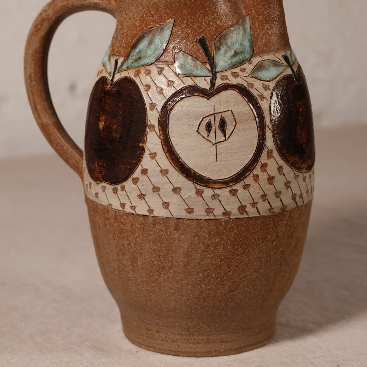 A handmade ceramic pitcher with a rounded brown body, a curved handle, and a decorative band featuring painted apple motifs and a geometric pattern.